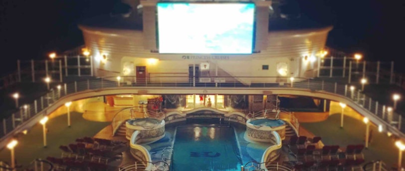 A luxury cruise ship deck featuring a pool surrounded by lounge chairs and warm ambient lighting. A large screen overlooks the area, showing an image against a dark night sky.