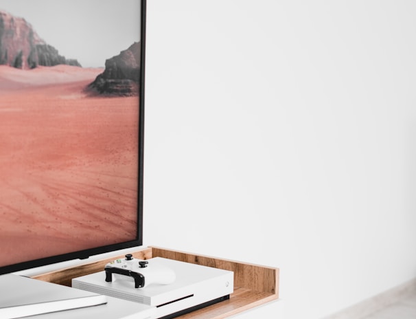 A modern interior setting featuring a white entertainment console with wooden accents. On the console, there is a gaming console with a controller on top. A large framed picture of a desert landscape with red sand and rocky formations is positioned next to the console.