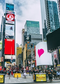 A bustling urban scene with towering skyscrapers and numerous digital billboards showcasing advertisements and entertainment content. A large crowd of people is visible, indicating a busy environment. Bright colors and diverse signage add to the vibrant atmosphere.