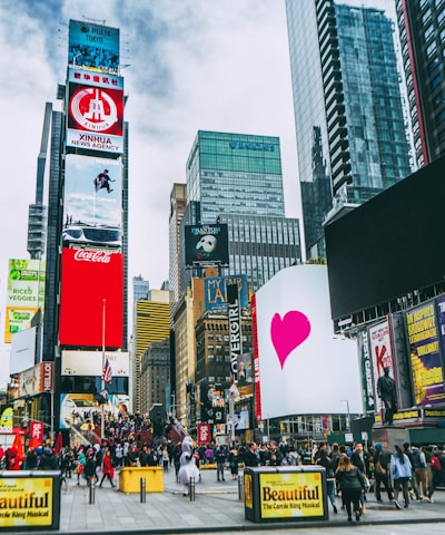 A bustling urban scene with towering skyscrapers and numerous digital billboards showcasing advertisements and entertainment content. A large crowd of people is visible, indicating a busy environment. Bright colors and diverse signage add to the vibrant atmosphere.