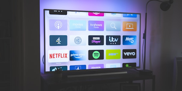 A flat-screen television is displayed in a dimly lit room, showcasing a variety of streaming service apps on its screen, such as Netflix, Prime Video, and Spotify. The TV is positioned on a black stand beside a tall floor lamp, casting a soft ambient light on the surrounding area.