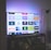 A flat-screen television is displaying a variety of streaming app icons, including Netflix, Prime Video, Spotify, and ITV Hub among others. The TV is placed on a media console with a small potted plant beside it. A tall lamp stands to the right, and a faint, ambient light casts a purple hue on the wall above the TV.