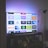 A flat-screen television is displaying a variety of streaming app icons, including Netflix, Prime Video, Spotify, and ITV Hub among others. The TV is placed on a media console with a small potted plant beside it. A tall lamp stands to the right, and a faint, ambient light casts a purple hue on the wall above the TV.