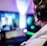 A person wearing headphones is sitting in front of a computer setup with multiple monitors displaying colorful, blurred graphics. The headphones have a distinctive logo illuminated in green. The ambient lighting and screen glow add to the immersive gaming environment.