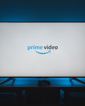 A television screen displays the Prime Video logo, situated on an entertainment unit in a dimly lit room. The room is illuminated with a subtle blue backlight, creating a calming atmosphere. Two small decorative items, resembling mushrooms, are placed on either side of the unit.