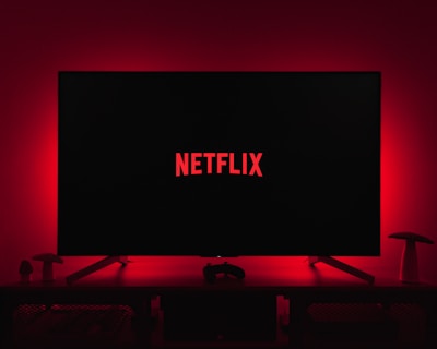 A dark room featuring a television screen displaying the word 'Netflix' in red against a black background. The screen is centered on a console table, with a video game controller placed in front. The room is dimly lit by red ambient lighting, creating a cozy atmosphere.
