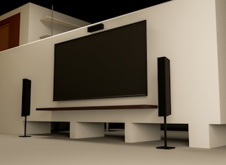 A modern home entertainment setup featuring a large flat-screen TV mounted on a light-colored wall. Two tall black speakers stand on either side of the TV. The room has a minimalist design with a clean, uncluttered look. Shelving and a wooden door are visible in the background.