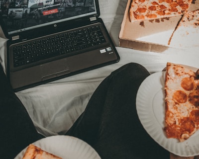 A cozy setting with a laptop displaying a streaming service interface nearby, alongside a pizza box holding a partially eaten pizza. Two slices of pizza are on paper plates held by someone, suggesting a casual movie night atmosphere.